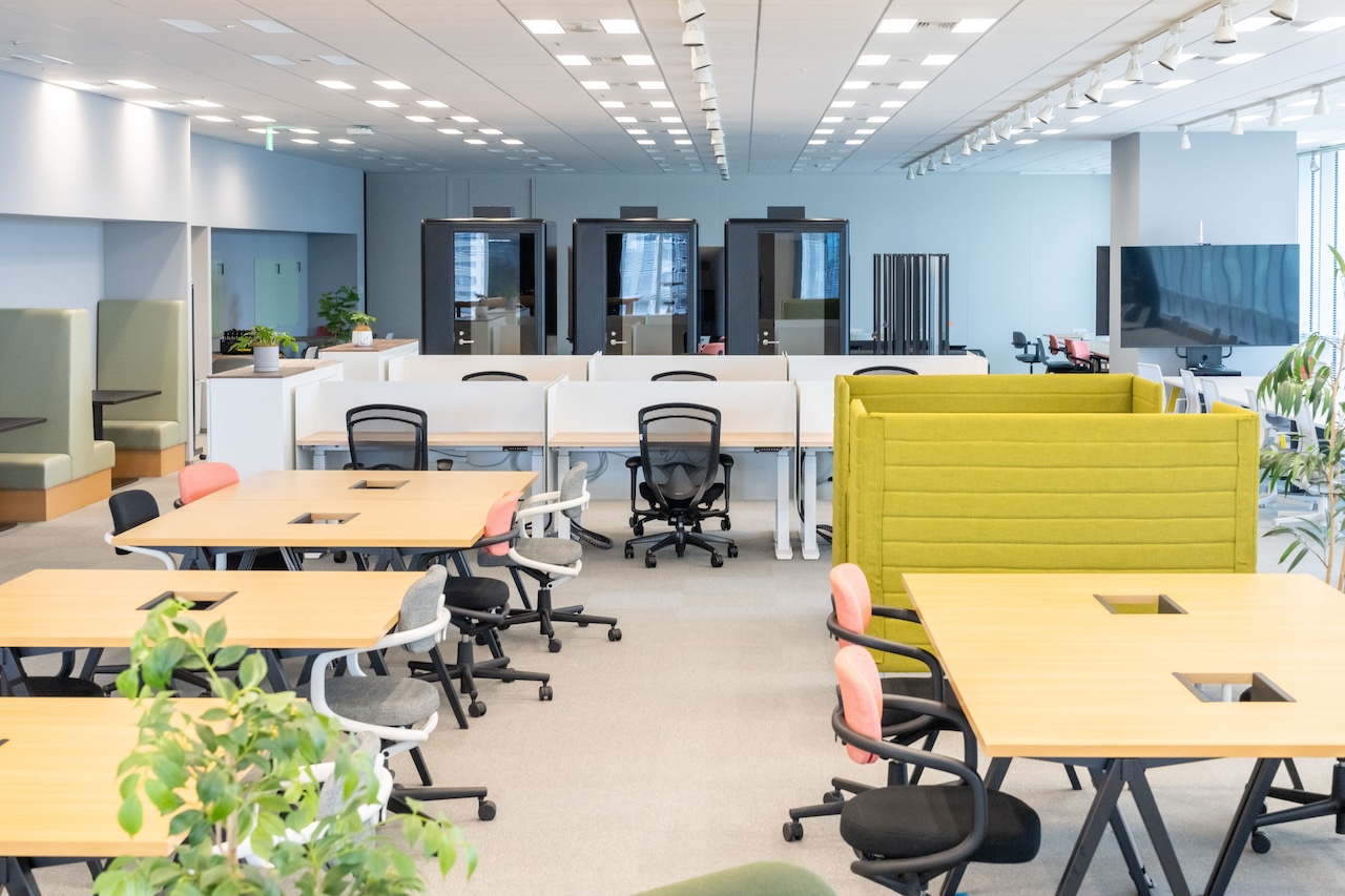 The spacious office room is furnished with a variety of desk and chair designs. Natural light pours in, accented by houseplants.