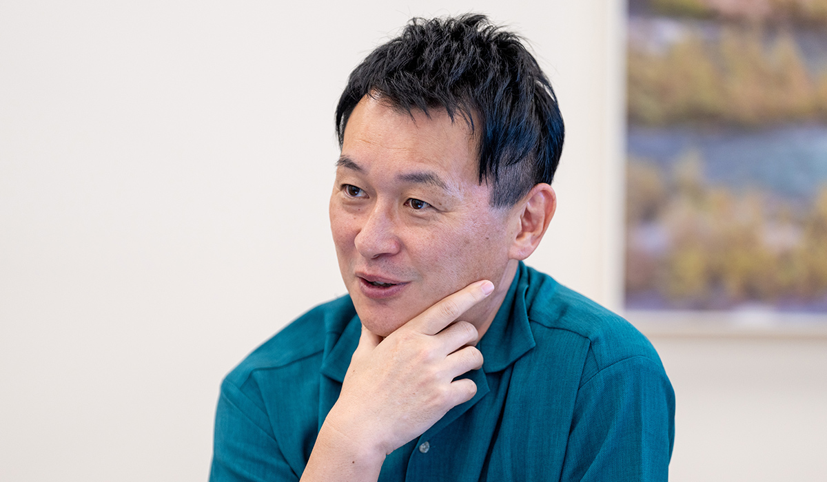 緑のシャツを着た坂上さんが、手をあごに当てているポーズをしています。